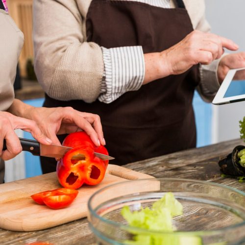 close-up-daughter-standing-with-her-mother-holding-digital-tablet-cutting-bell-pepper-with-knife_23-2148041513